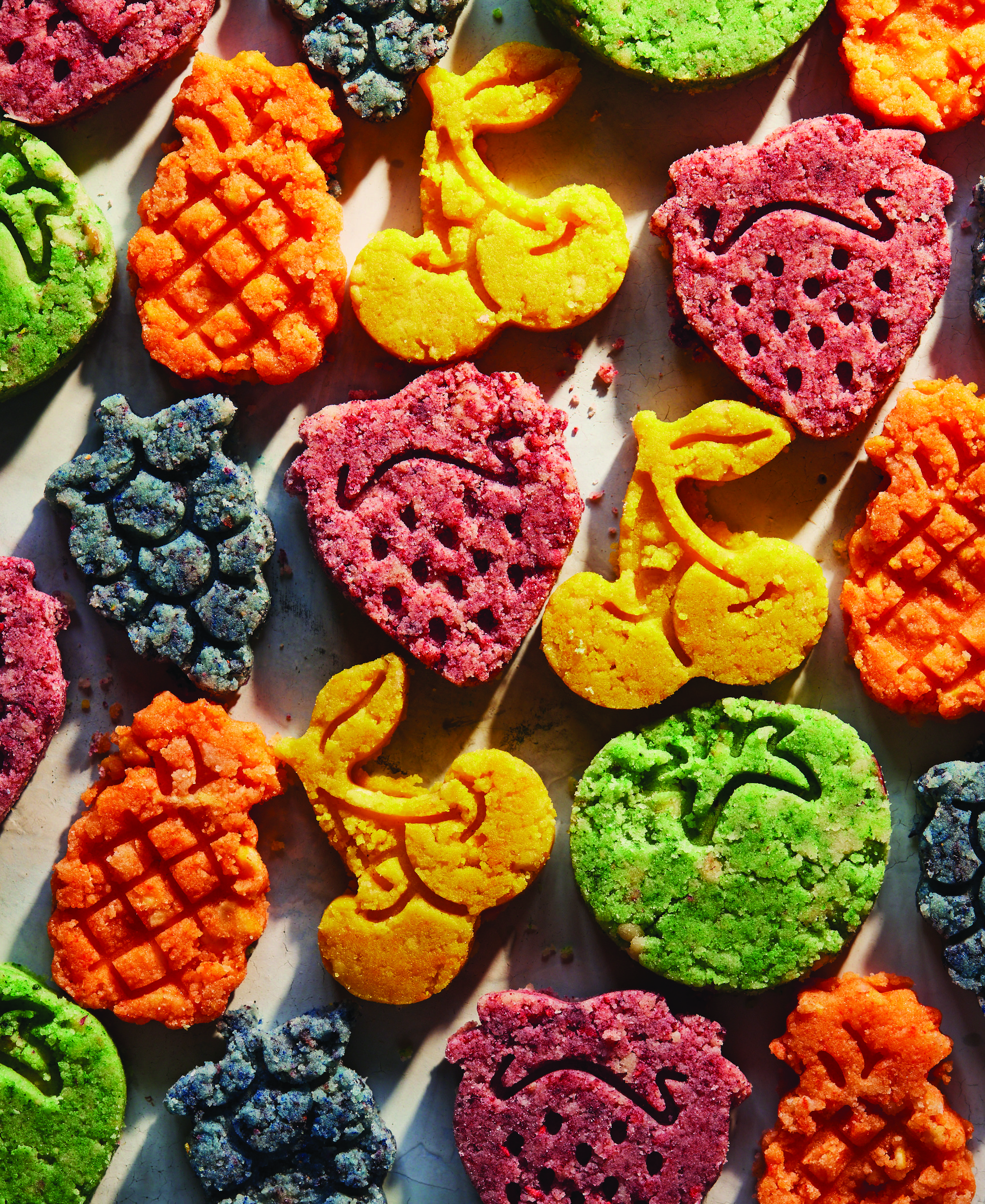 A colorful selection of rainbow polvoron. The cookies are shaped like cherries, pineapple, grapes, strawberries, and apples.