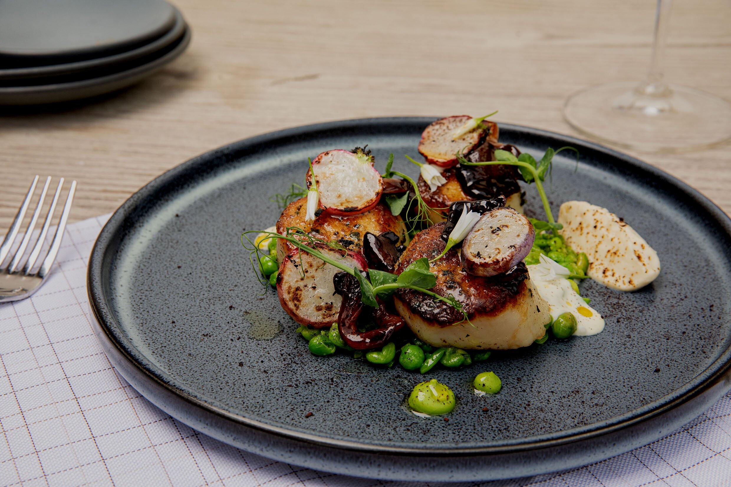 Seared scallops on a black plate surrounded by seared radishes, peas, and pea greens.