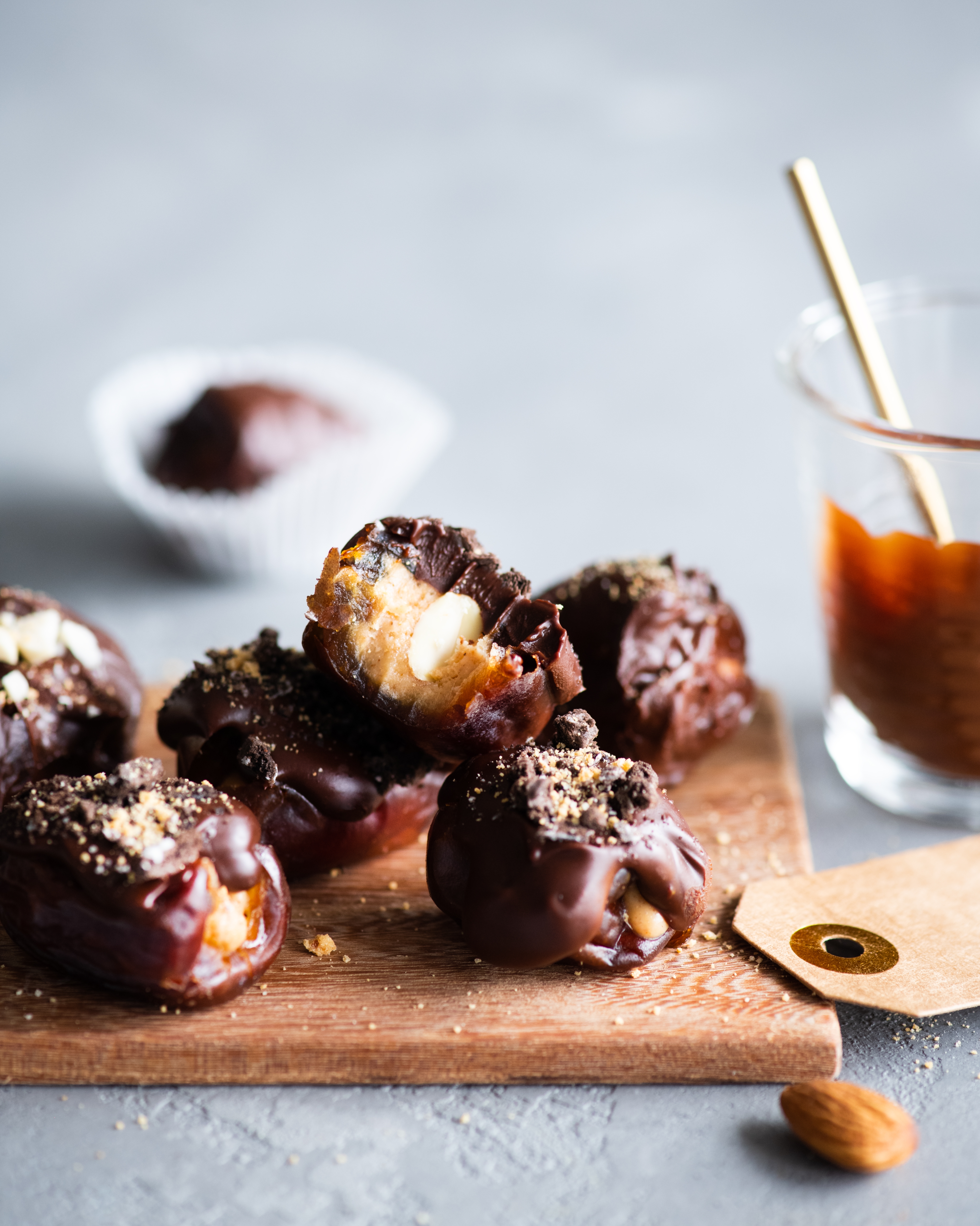 A handful of purple dates topped with chocolate and crushed nuts on a cutting board, next to a brown paper gift tag and a glass jar of peanut butter.