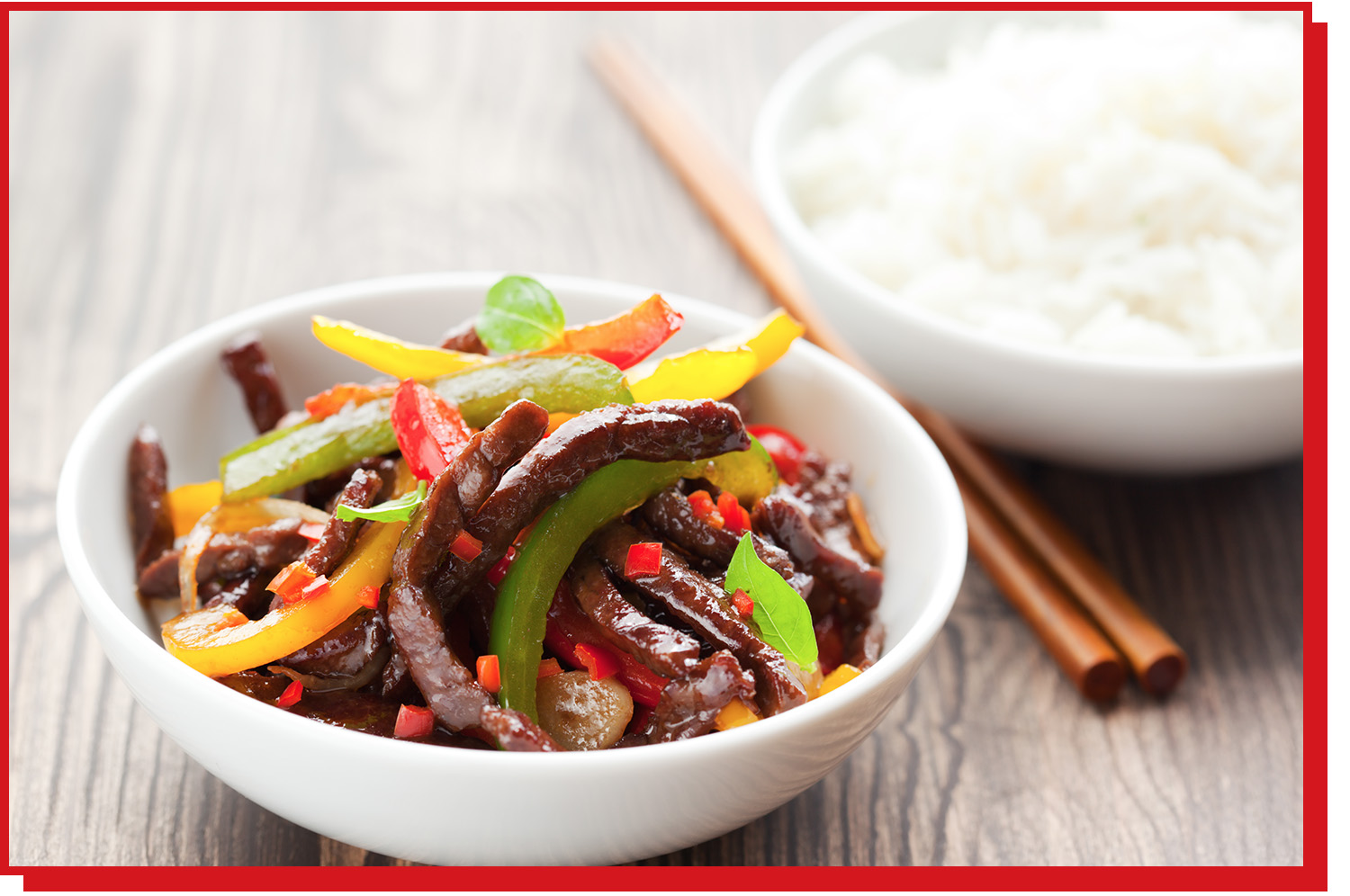 A bowl of beef and vegetable stir fry sits on a tabletop, with chopsticks and rice.