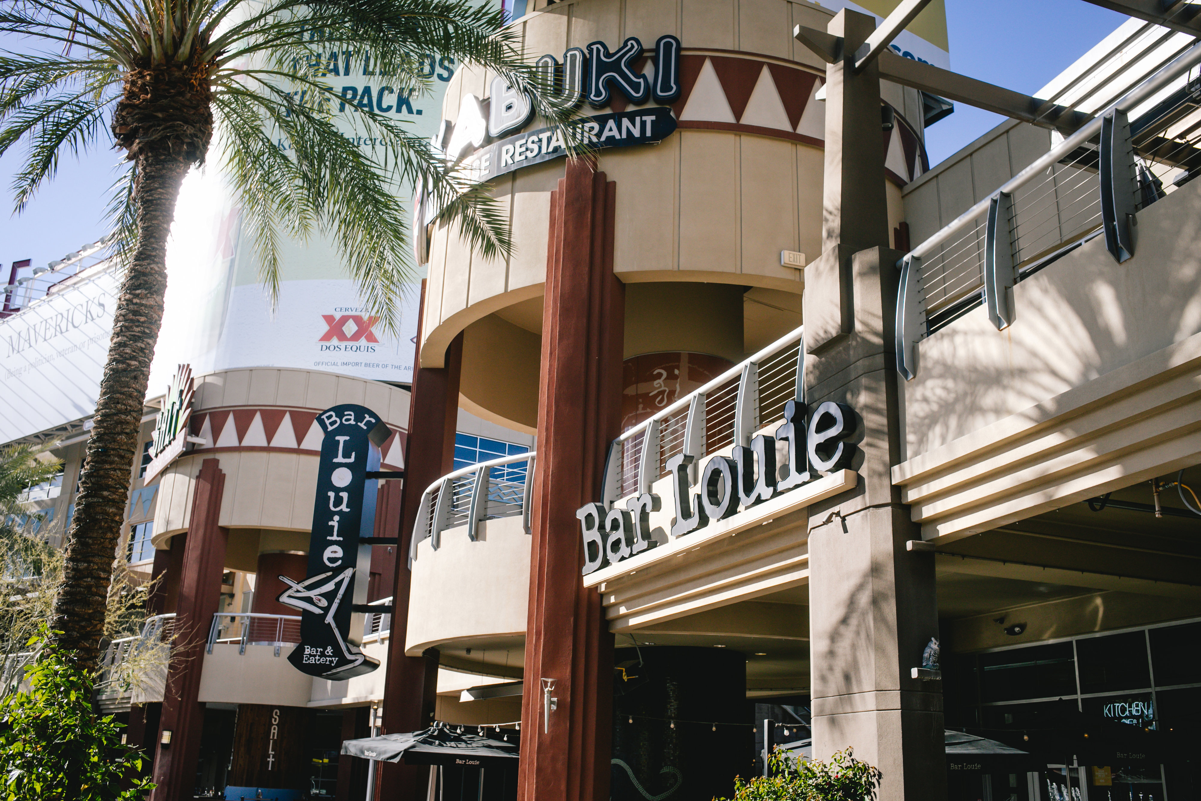 The exterior of a a building with signs for Kabuki Restaurant and Bar Louie is surrounded by palm trees on a sunny day. 